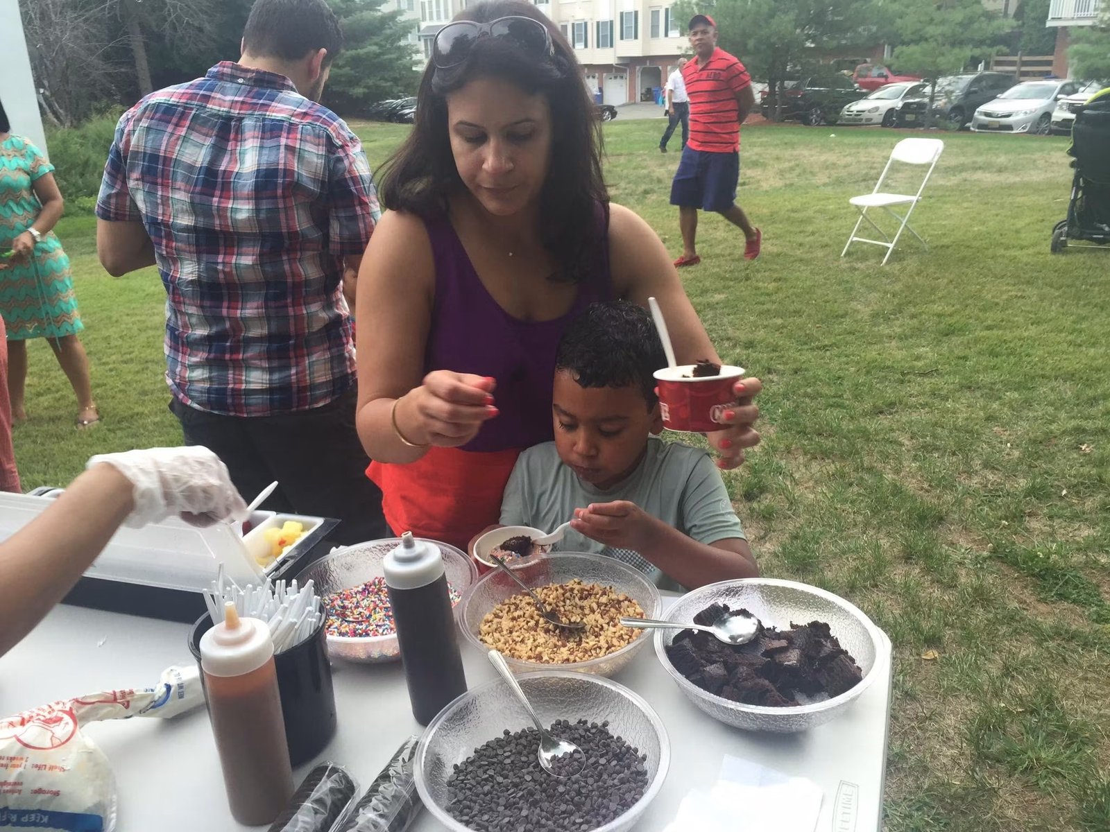 A parent and child at an outdoor Cold Stone toppings table on the grass, with mix-ins ready to serve
