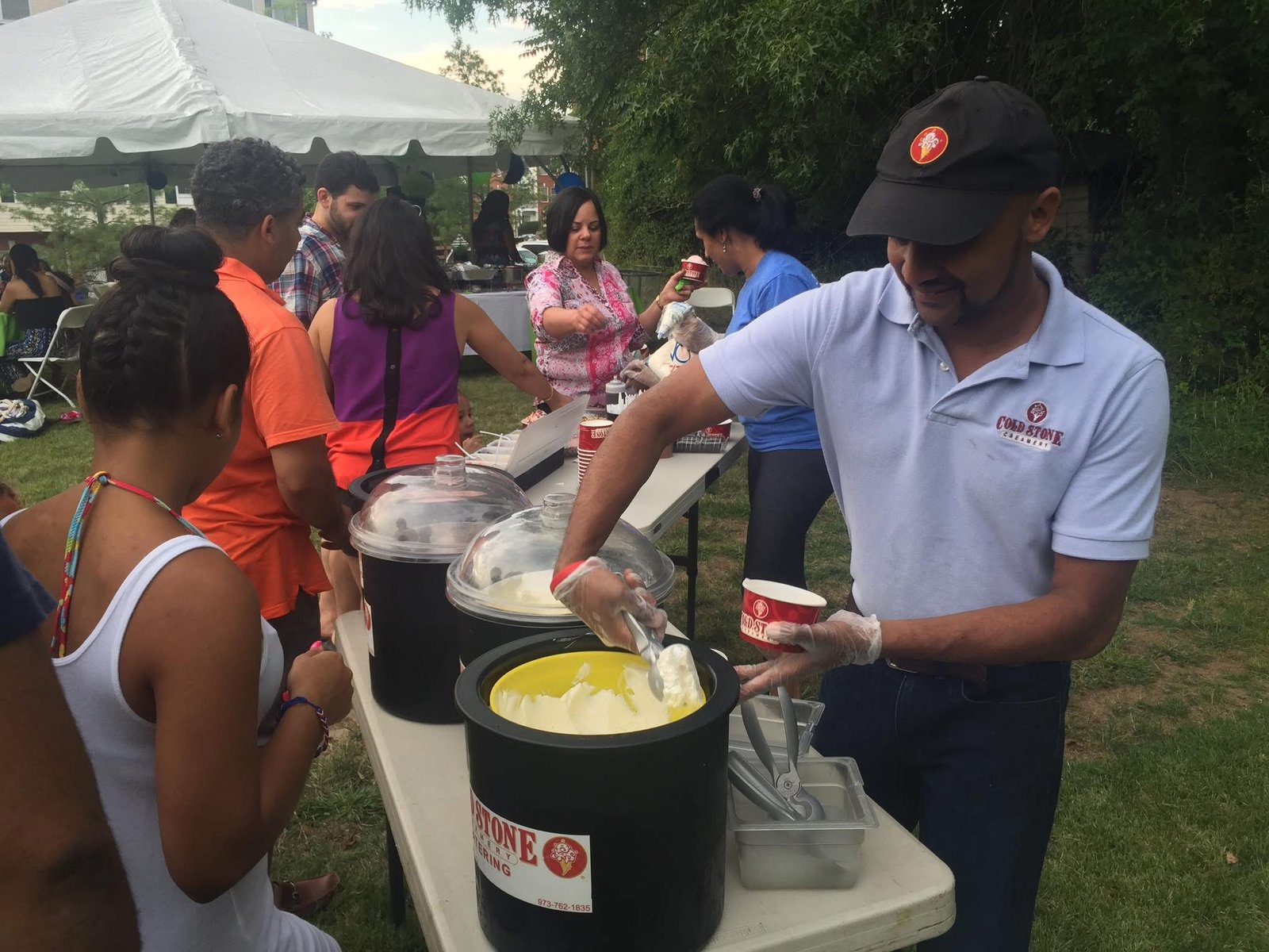 A Cold Stone team member scooping ice cream from catering tubs to serve guests at an outdoor tented event